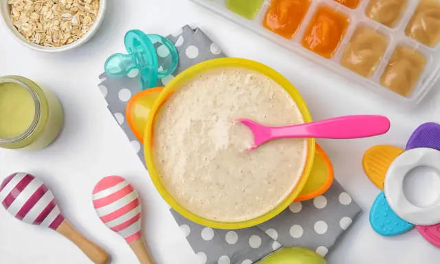 Un tazón de papilla de cereales con una cuchara rosa, junto a avena, purés de frutas y juguetes para bebés.