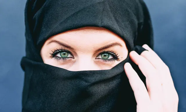 Ein schwarzes Kopftuch umrahmt ein Gesicht, dessen grüne Augen durch den Stoff blicken. Die Bedeutung von Kopftuchfarben ist vielfältig.