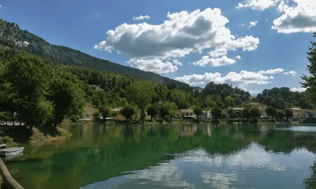 Il lago Sirino, con le sue acque verdi e calme, riflette le case e le colline boscose sotto un cielo azzurro con nuvole bianche.