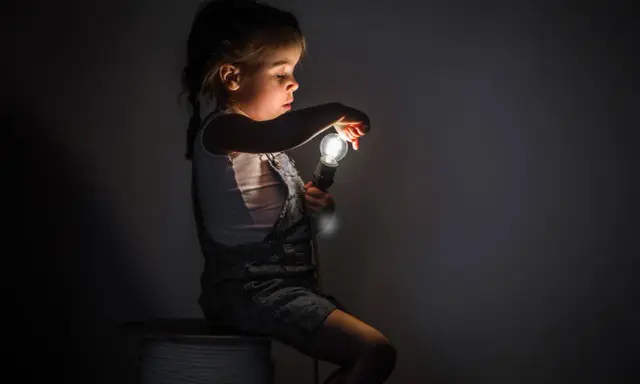 Niña con miedo a la oscuridad, sentada en un rollo de cable, sosteniendo una bombilla encendida.
