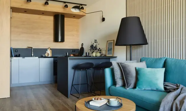 Nowoczesny salon z aneksem kuchennym 22m2. Turkusowa kanapa, czarne hokery przy wyspie, drewniane akcenty i stylowe oświetlenie tworzą przytulną przestrzeń.