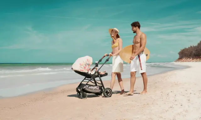 Para spaceruje po plaży z wózkiem Kinderkraft Askoy. Mężczyzna niesie deskę surfingową.