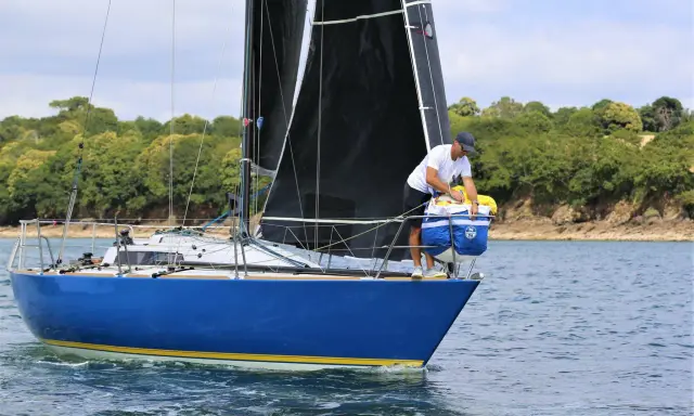 Un homme prépare le bateau, une voile noire déployée, sur fond de côte boisée. Il semble être le **half toner** de l'équipage.