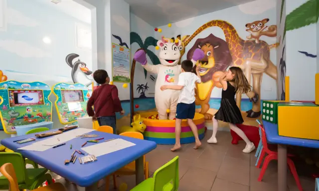 Bambini giocano con un personaggio in costume da ippopotamo in una stanza colorata, un'area giochi per famiglie al Mokambo Shore Easy Family Hotel.