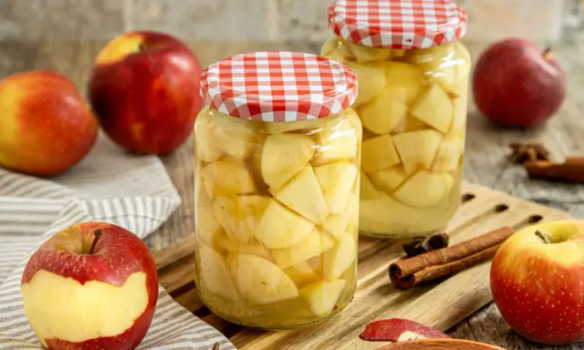 Zwei Gläser mit Apfelstücken, bereit zum Apfel einkochen. Frische Äpfel, Zimtstangen und Sternanis liegen daneben.