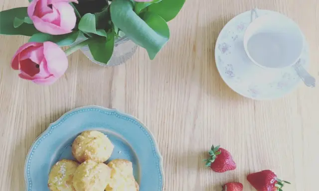 Chouquettes traditionnelles sur une assiette bleue, accompagnées de fraises et d'un bouquet de tulipes roses. Parfait pour une pause gourmande.