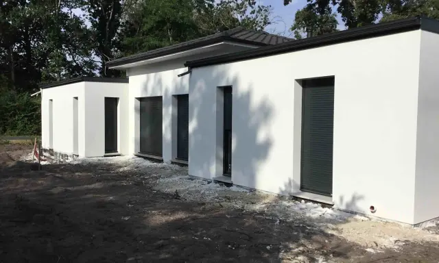 Maison moderne avec toit plat, plan de 150m2. Construction blanche aux lignes épurées, entourée d'arbres.