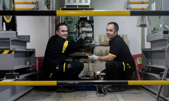 Técnicos realizan mantenimiento de elevadores en un taller, revisando la electrónica y cableado.