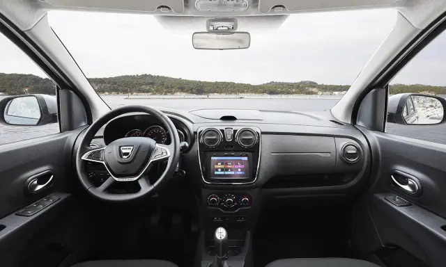 Interior del Dacia Lodgy con volante, pantalla táctil y palanca de cambios. El paisaje marino se ve a través del parabrisas.