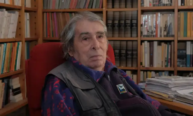 Ferdinando Camon, con i suoi capelli grigi e lo sguardo pensieroso, siede tra scaffali pieni di libri, un ritratto di un intellettuale immerso nel suo mondo.