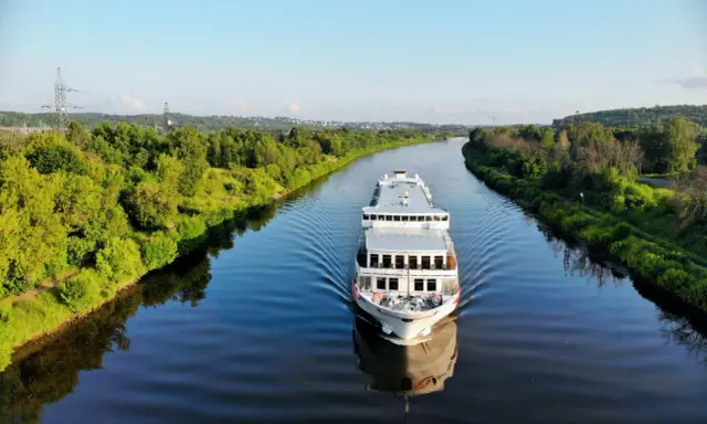 Nave da crociera solca un fiume sereno, con rive alberate che ricordano la bellezza naturale vicina alla capitale Russia.