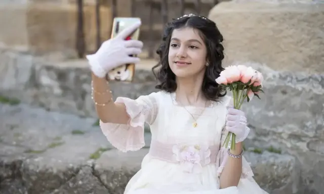 Dziewczynka w sukni komunijnej robi selfie z bukietem kwiatów. To ważny moment, przygotowania do pierwszej komunii świętej krok po kroku.