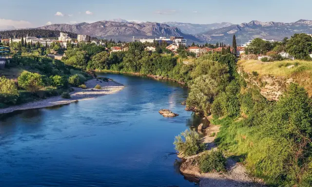 Malowniczy widok na rzekę i miasto Podgorica. W oddali góry, a na pierwszym planie bujna zieleń i kamieniste brzegi. Atrakcje Podgoricy w pełnej krasie.