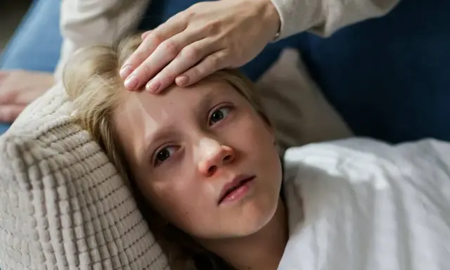 Niña con fiebre, su frente tocada por una mano. Posiblemente un caso de mononucleosis en niños.