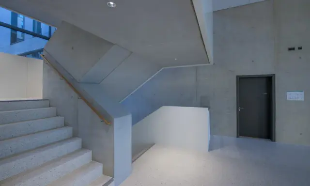 Moderne Treppe mit Sichtbeton-Wänden, die durch die Schalung eine interessante Textur erhalten. Ein dunkler Türrahmen und ein Holzhandschuh ergänzen das Bild.