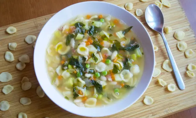 Un reconfortante plato de sopa minestrone, lleno de pasta orecchiette, verduras y frijoles, servido en un tazón blanco sobre una tabla de madera.