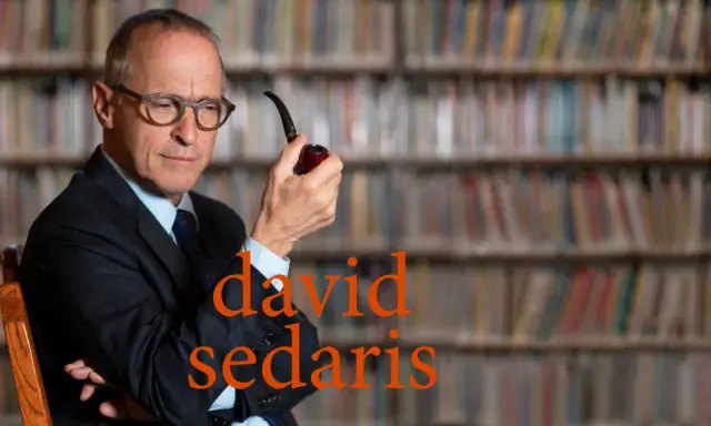 David Sedaris, con pipa in mano, seduto tra scaffali pieni di libri, riflette pensieroso.