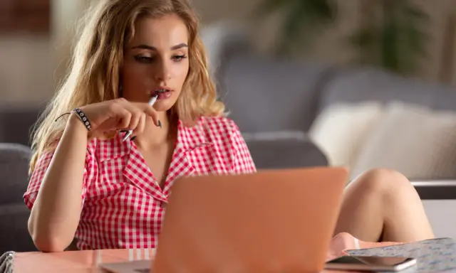 Młoda kobieta w kratkę zastanawia się, jak zacząć rozmowę przez internet, trzymając długopis przy ustach.
