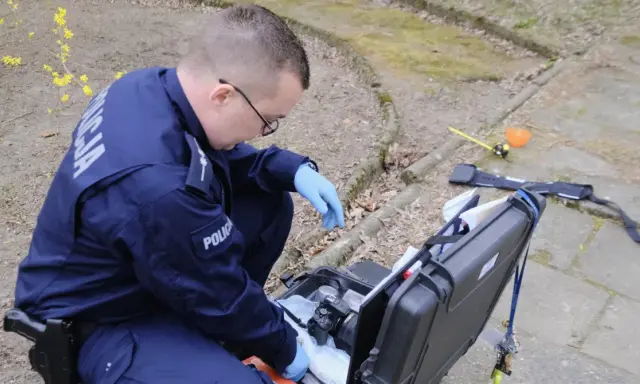 Policjant w niebieskim mundurze z napisem "POLICJA" klęczy przy otwartej walizce z aparatem i sprzętem kryminalistycznym.