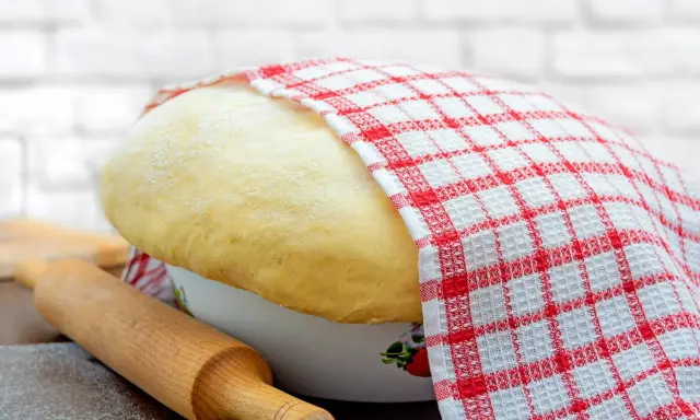 Wyrośnięte ciasto drożdżowe, przykryte ręcznikiem w kratkę, czeka na swój przepis babci. Obok wałek i mąka.