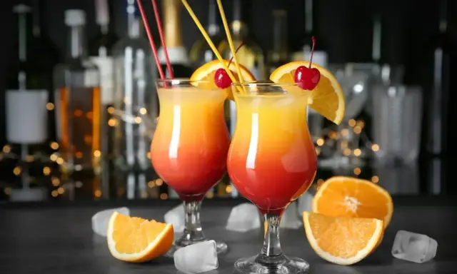 Dwa drinki z whisky ze spritem, ozdobione pomarańczą i wiśnią, na tle barowych butelek.