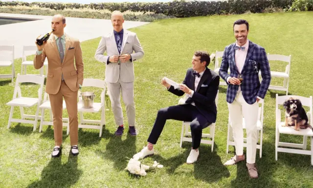 Four men in stylish garden formal wedding attire for male guests pose on a lawn. One drinks champagne, another adjusts his suit, one sits with a bouquet, and the last holds a drink with a dog nearby.