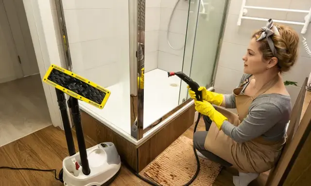 Kobieta w żółtych rękawiczkach czyści prysznic parownicą, walcząc z pleśnią w łazience. Obok stoi urządzenie i nakładka.
