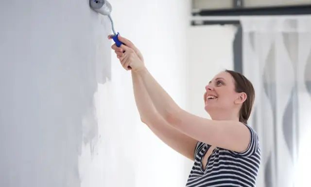 Une femme souriante apprend comment peindre un mur avec un rouleau. Elle applique une couche de peinture grise sur un mur blanc.