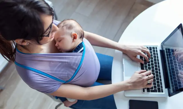 Kobieta w chuście nosi śpiące dziecko i pracuje na laptopie. Czy na macierzyńskim można pracować? Tak, to dowód.