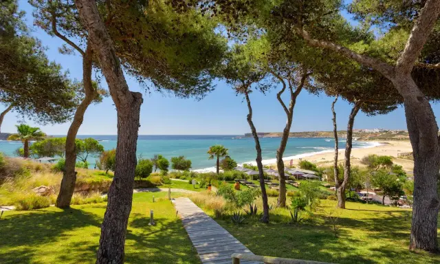 Holzweg führt zu einem sonnigen Strand in Pera, Portugal. Pinienbäume säumen den Weg zum türkisfarbenen Meer.