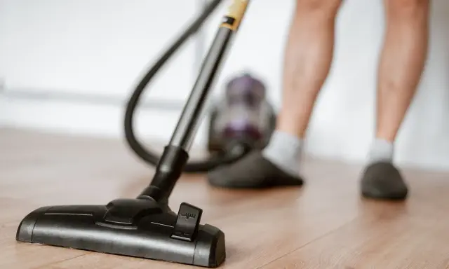 Odkurzacz na drewnianej podłodze, część nóg w kapciach i skarpetkach. To jeden z obowiązków domowych po angielsku – "vacuuming".
