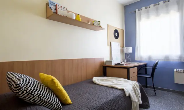 Habitación de estudiante en la carlos v residencia granada. Cama con cojines, escritorio con lámpara y estantería con libros.