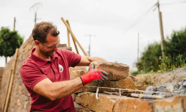 Un maçon, ganté de rouge, pose une pierre pour construire un mur en Corse.