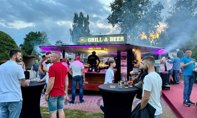 Ludzie przy stoisku z grillem i piwem, z dymem unoszącym się w powietrzu.