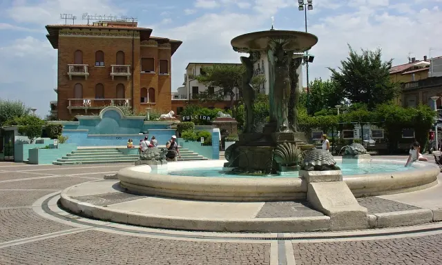 Fontana con statue e tartarughe in una piazza di Cattolica centro, con edifici storici e persone che si godono il sole.