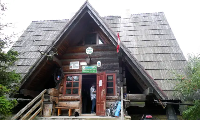 Drewniana bacówka PTTK pod Małą Rawką z otwartymi drzwiami, gdzie stoi mężczyzna. Obok stoją rowery i plecaki.