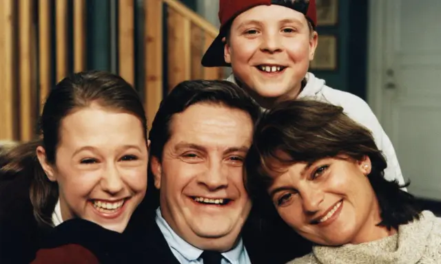 En glad familj, som hämtad ur svenska komedier, ler mot kameran. Pappa, mamma, dotter och son.