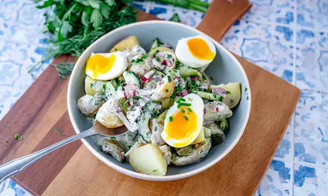 Schüssel mit cremigem Kartoffelsalat mit Gurken, Zwiebeln und gekochten Eiern, garniert mit frischen Kräutern.