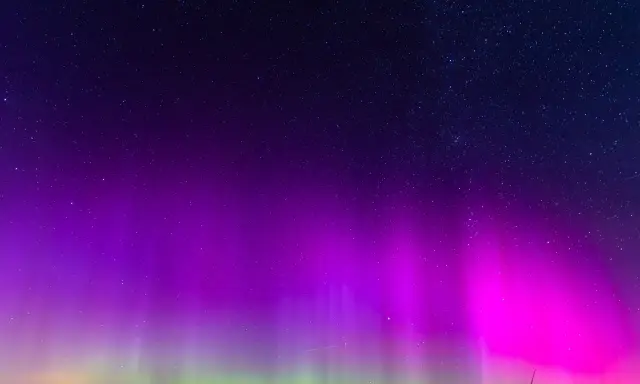 Nocne niebo rozświetlone fioletową i zieloną zorzą polarną, kiedy jest zorza polarna. Gwiazdy migoczą nad ciemnym horyzontem.