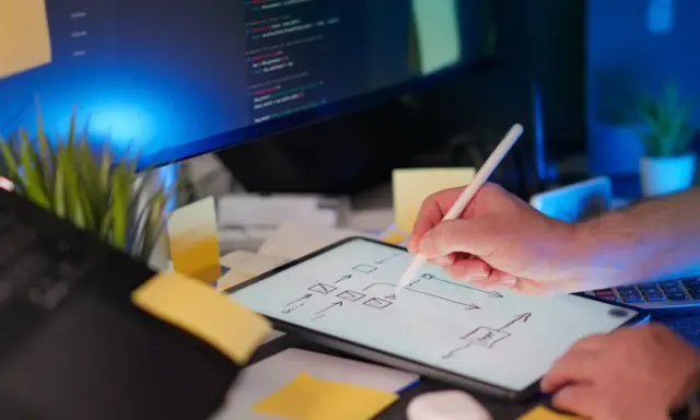 Programista szkicuje schemat blokowy na tablecie, planując implementację **typescript abstract class**. Na ekranie komputera widać kod.
