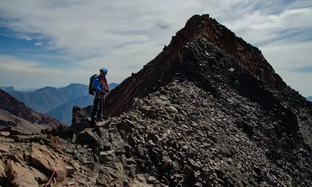 Alpinista na grani, zdobywający najwyższy szczyt Gruzji. W tle skaliste pasmo górskie i błękitne niebo.