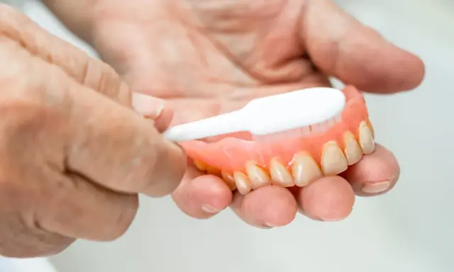 Dłonie czyszczą protezę zębową szczoteczką, pokazując jak usunąć kamień z plastiku.