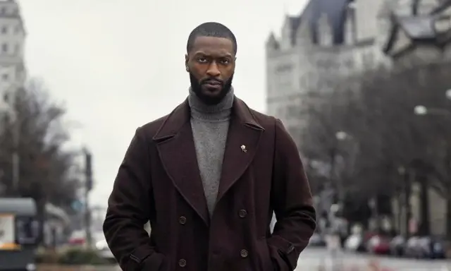 Alex Cross, un uomo con un cappotto scuro e un dolcevita grigio, guarda intensamente verso l'obiettivo.