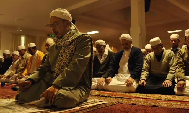 Männer in weißen Turbanten und traditioneller Kleidung knien auf Gebetsteppichen und verrichten das Taraweeh-Gebet.