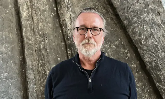 Thomas Olsson, en man med glasögon och grått skägg, står framför en grov, stenliknande vägg.