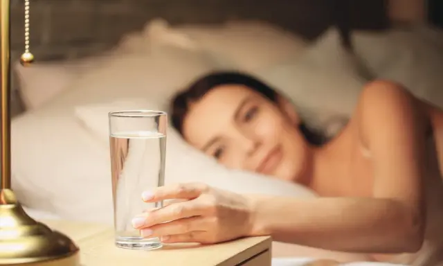 Une main attrape un verre d'eau posé à droite sur une table de chevet, une femme se réveille en arrière-plan.