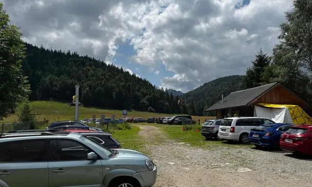 Parking z samochodami przy górskim schronisku, w tle las i chmury.