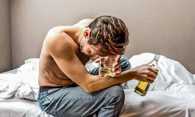 Mężczyzna siedzi na łóżku, trzymając szklankę z alkoholem i butelkę.