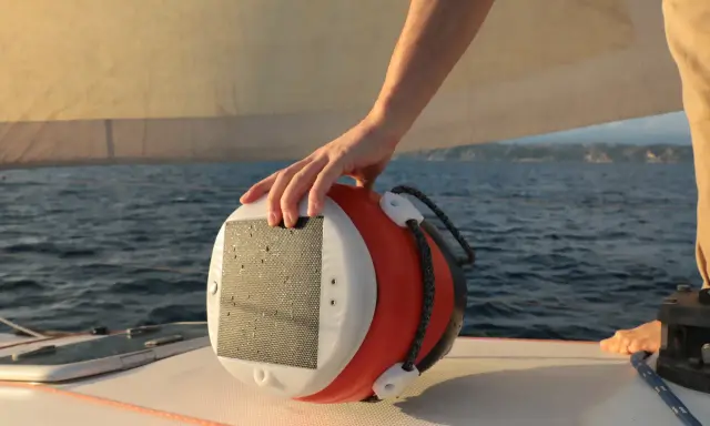 Main d'un marin tenant une bouée orange et blanche, un câble y est accroché. Le bateau navigue sur une mer bleue sous un ciel clair.