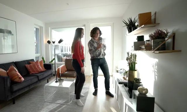 Två personer diskuterar något i ett modernt vardagsrum med trendig inredning, mörk soffa och hyllor med växter.